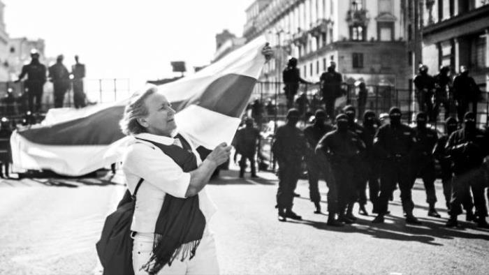 Osamocená žena v bílém drží tradiční běloruskou vlajku - bíločervenobílou bikolóru. Vlajka a vlasy ženy vlají ve vzduchu. Za ženou je skupina vládních jednotek - ozbrojenců v černém.